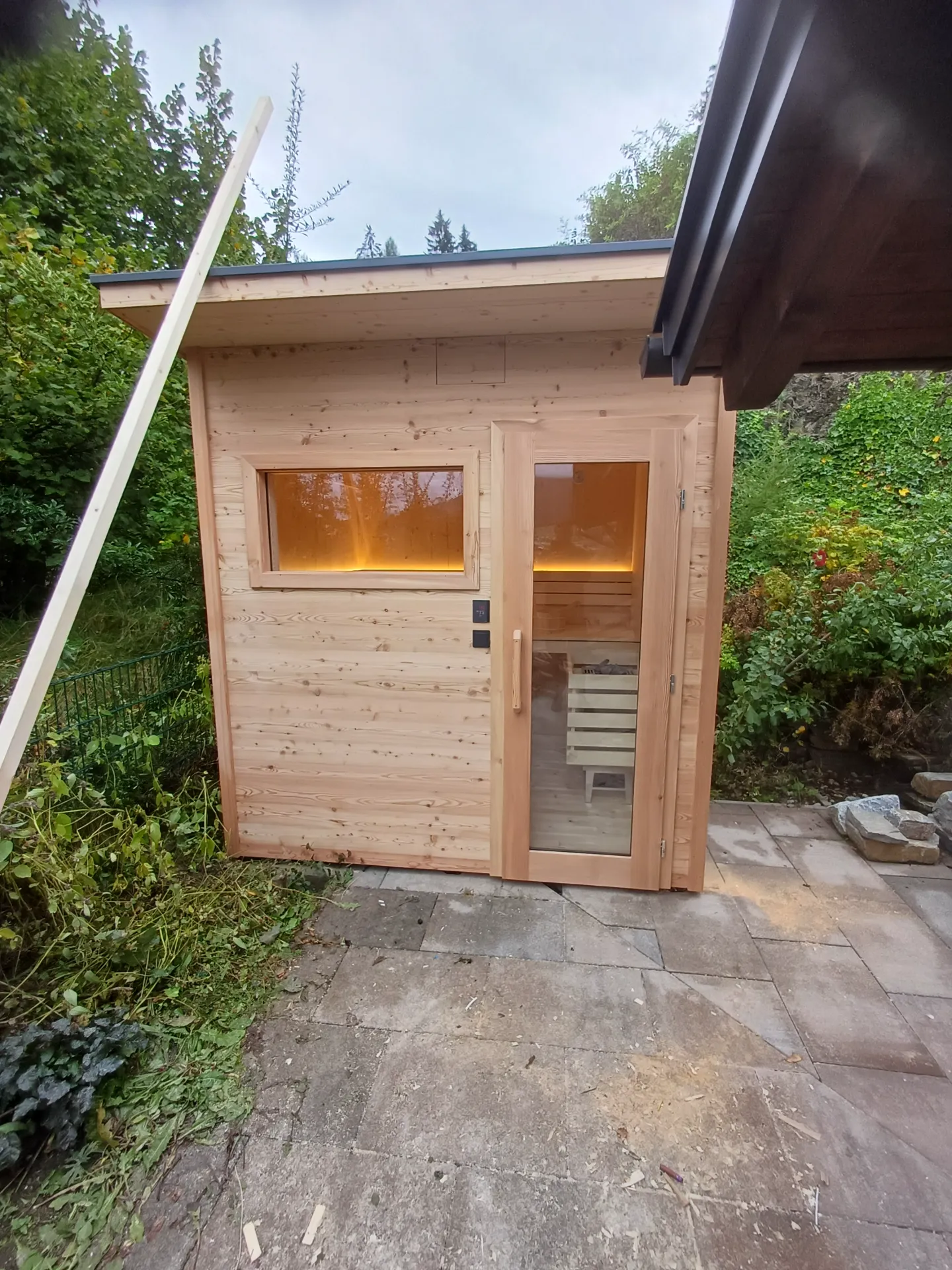 Neue Außensauna aus Lärchenholz mit Flachdach, Glastür und beleuchtetem Fenster im Garten