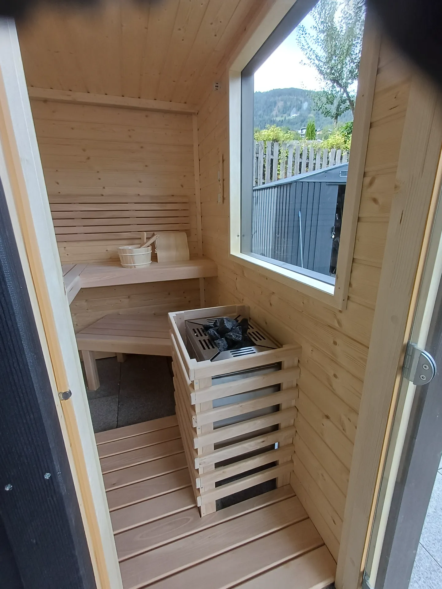 Innenraum einer Außensauna mit Saunaofen, Holzbänken und Fenster mit Bergblick